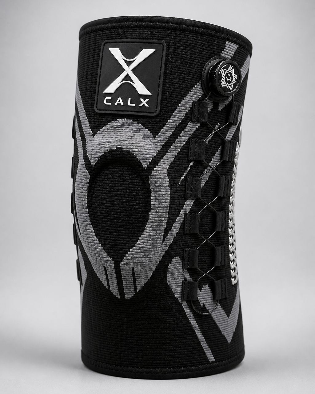 A black and gray CALX knee support sleeve with a black logo patch on the front and a side adjustment dial, shown on a neutral gray background.